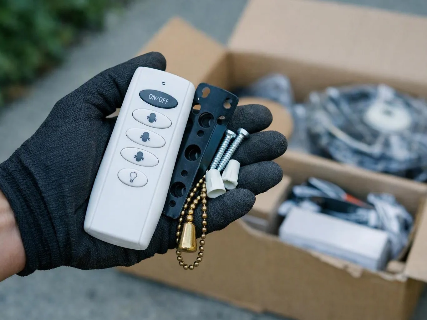 A gloved hand holds a white fan remote, mounting bracket, screws, and a pull chain in front of an open box