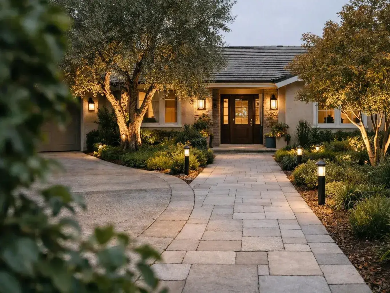 Warmly lit suburban house with stone walkway, trees, and garden lights at dusk