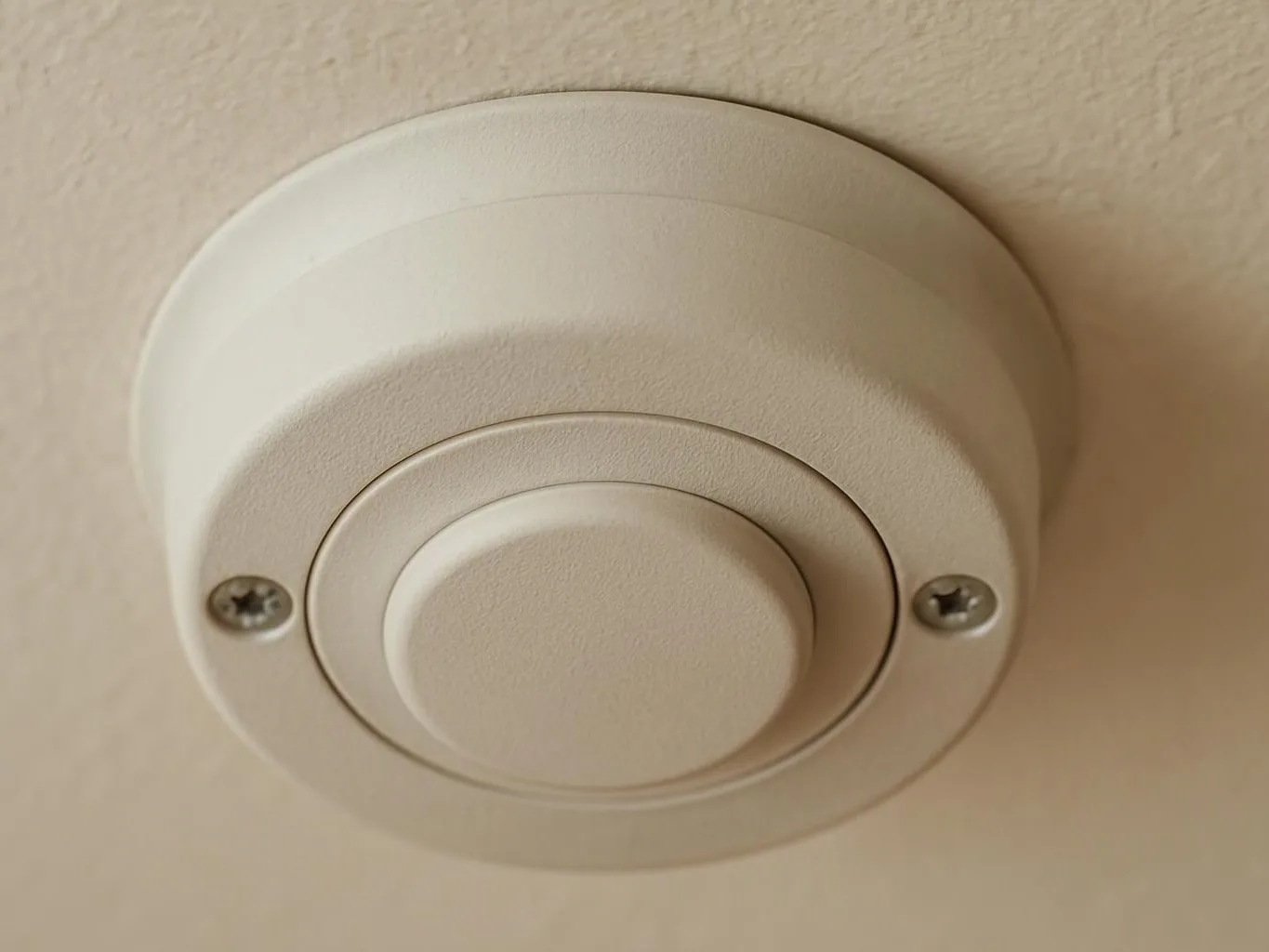 A round white ceiling smoke detector sits on a beige ceiling with two visible screws